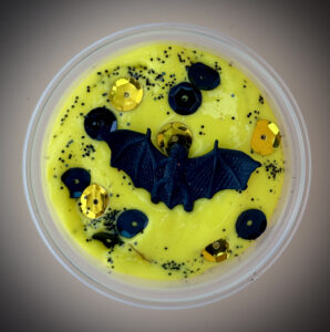 A vibrant yellow slime with a black bat toy and black glitter and black and gold sequins inside a clear plastic container.