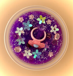A vibrant purple slime with a pink plastic flower toy ring and various plastic gems and flower sequins inside a clear plastic container.