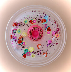 A white slime with a pink doughnut toy and various colorful fruit sequins and glitter inside a clear plastic container.