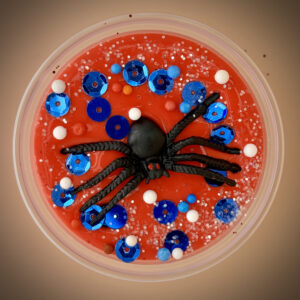 A red slime with a black spider toy and blue sequins and white balls inside a clear plastic container.