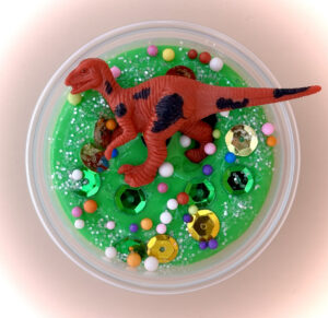 A plastic dinosaur toy standing in a bowl of green slime decorated with colorful beads and shiny sequins.