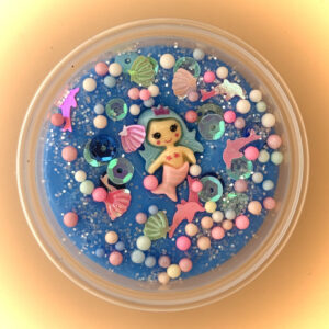 A blue slime with a colourful mermaid toy and various colourful beads and sequins inside a clear plastic container.